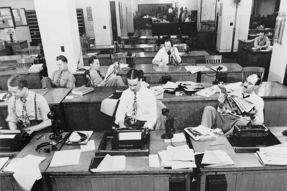 Interiör från en äldre tidningsredaktion där journalister skriver på skrivmaskin, talar i telefon och läser i tidningen